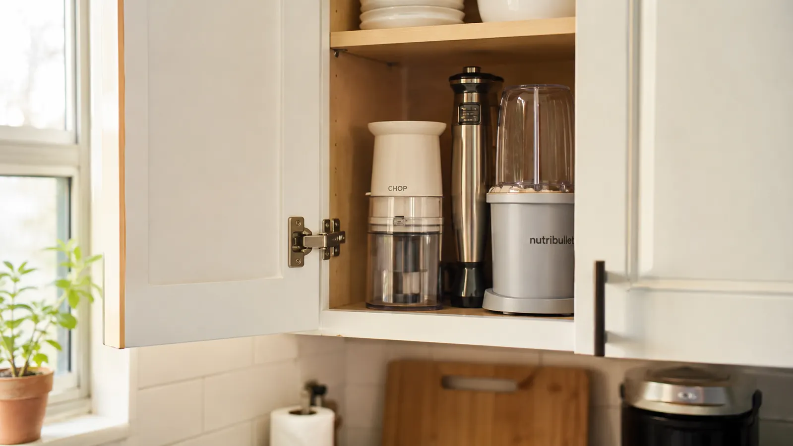 A small kitchen cabinet shelf with the chopper stored next to a few other compact tools, lid off, showing how little space it takes