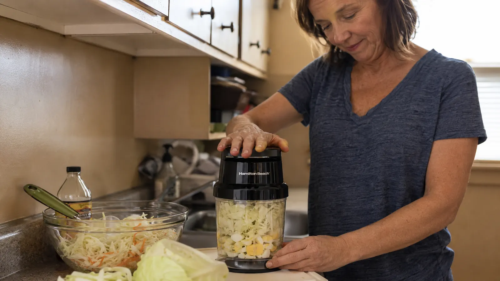Hand pressing down on the Hamilton Beach chopper lid while shredded cabbage and a hard-boiled egg sit in the clear bowl mid-chop