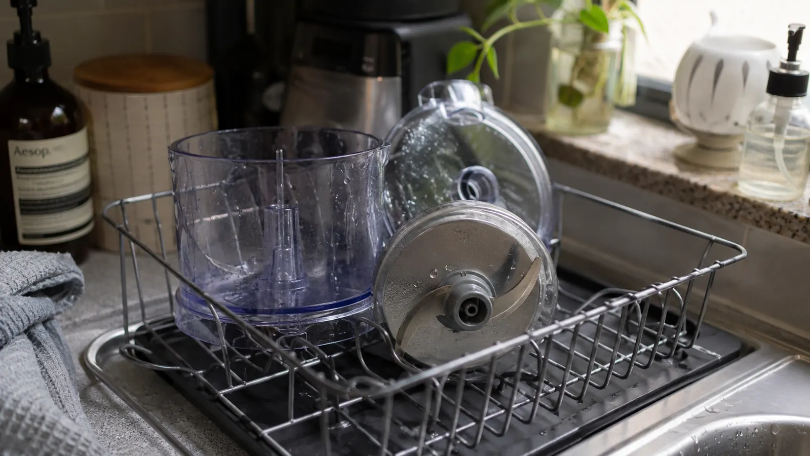 Small chopper bowl, lid, and blade sitting in a dish rack next to a sponge after a quick rinse