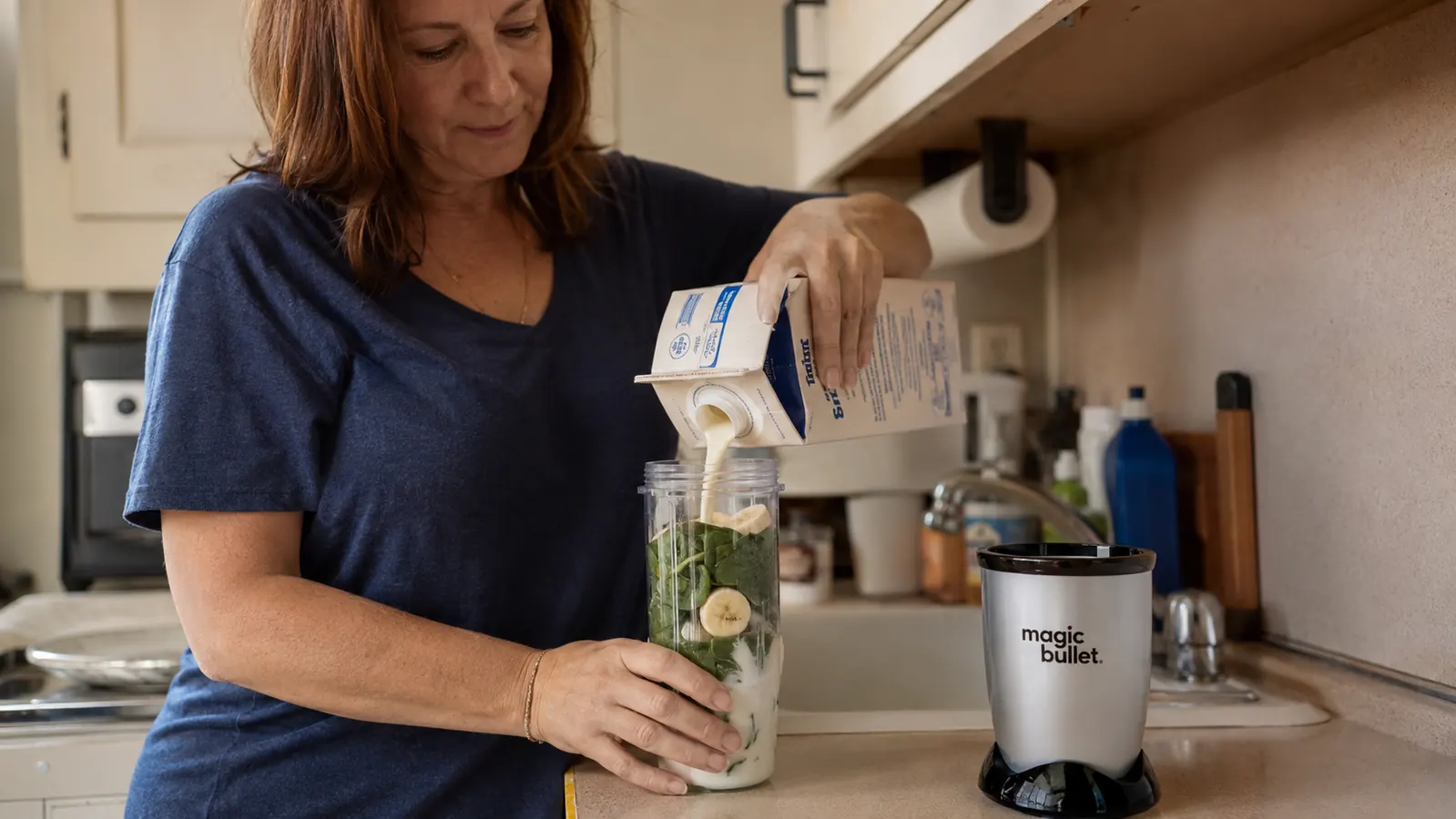 Hand pouring almond milk into a Magic Bullet cup already loaded with spinach and banana, up to the fill line printed on the cup