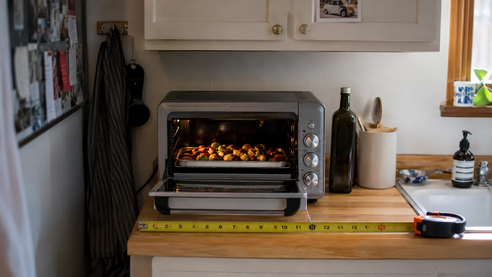 Small baking tray of vegetables sliding into the toaster oven next to a measuring tape showing the unit's width on the counter